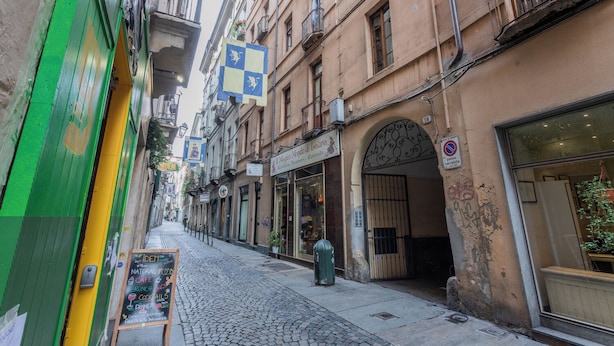 A narrow cobblestone street lined with buildings, including a café with outdoor seating and a store with a large window. - Barbaroux Apartments by Wonderful Italy (Turin)