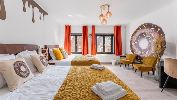 Habitación de hotel con dos camas, un ventanal con cortinas naranja y un cuadro de un donut, además de una gran decoración con forma de donut en la pared.