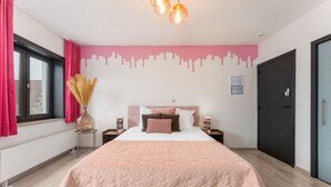 Une chambre à coucher avec un lit, une table de chevet, un vase, une fenêtre avec des rideaux et une porte.