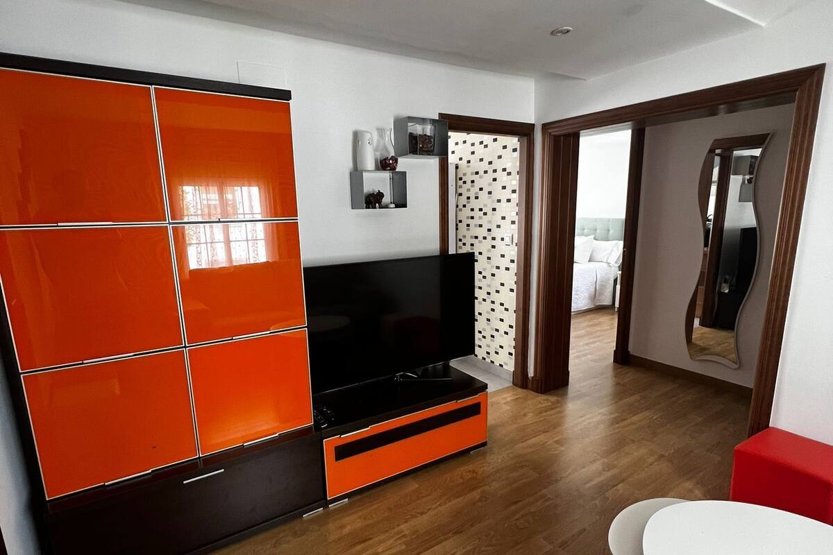 A modern living room with a large flat-screen TV, orange cabinets, a small white table, and a red ottoman.