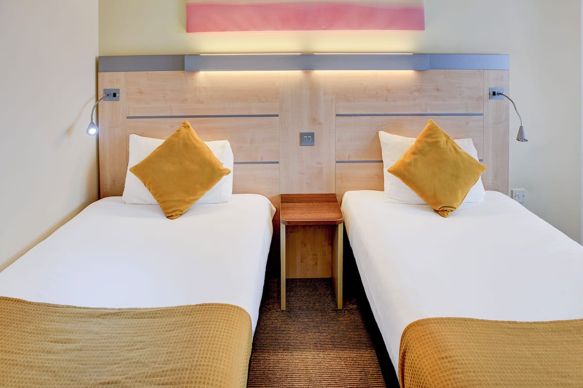 Quarto de hotel com duas camas de solteiro, cada uma com cabeceira de madeira, uma pequena mesa de cabeceira de madeira e luminária fixada na parede.