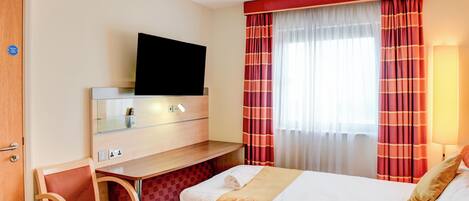 A hotel room with a bed, a desk with a flat-screen TV, a chair, and a window with curtains.