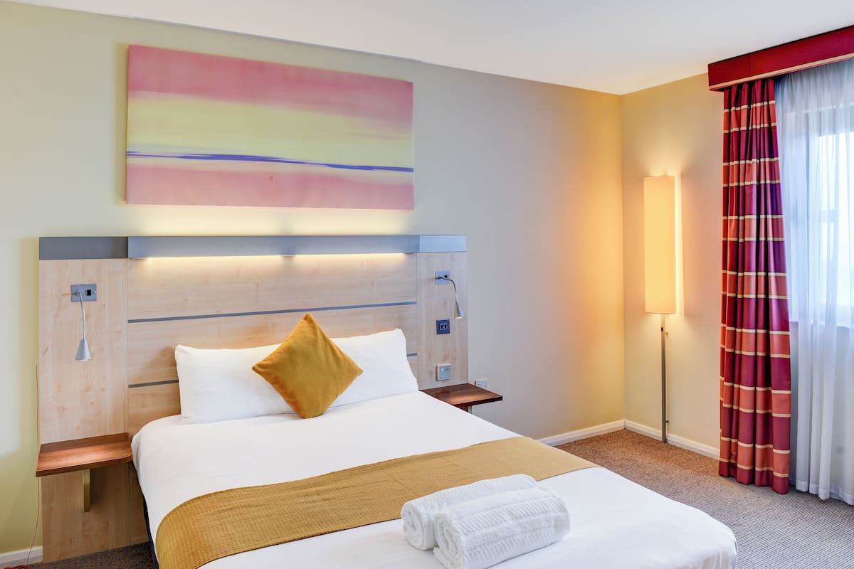 Quarto de hotel com cama bem arrumada, luminária de cabeceira, obra de arte na parede e janela com cortinas.