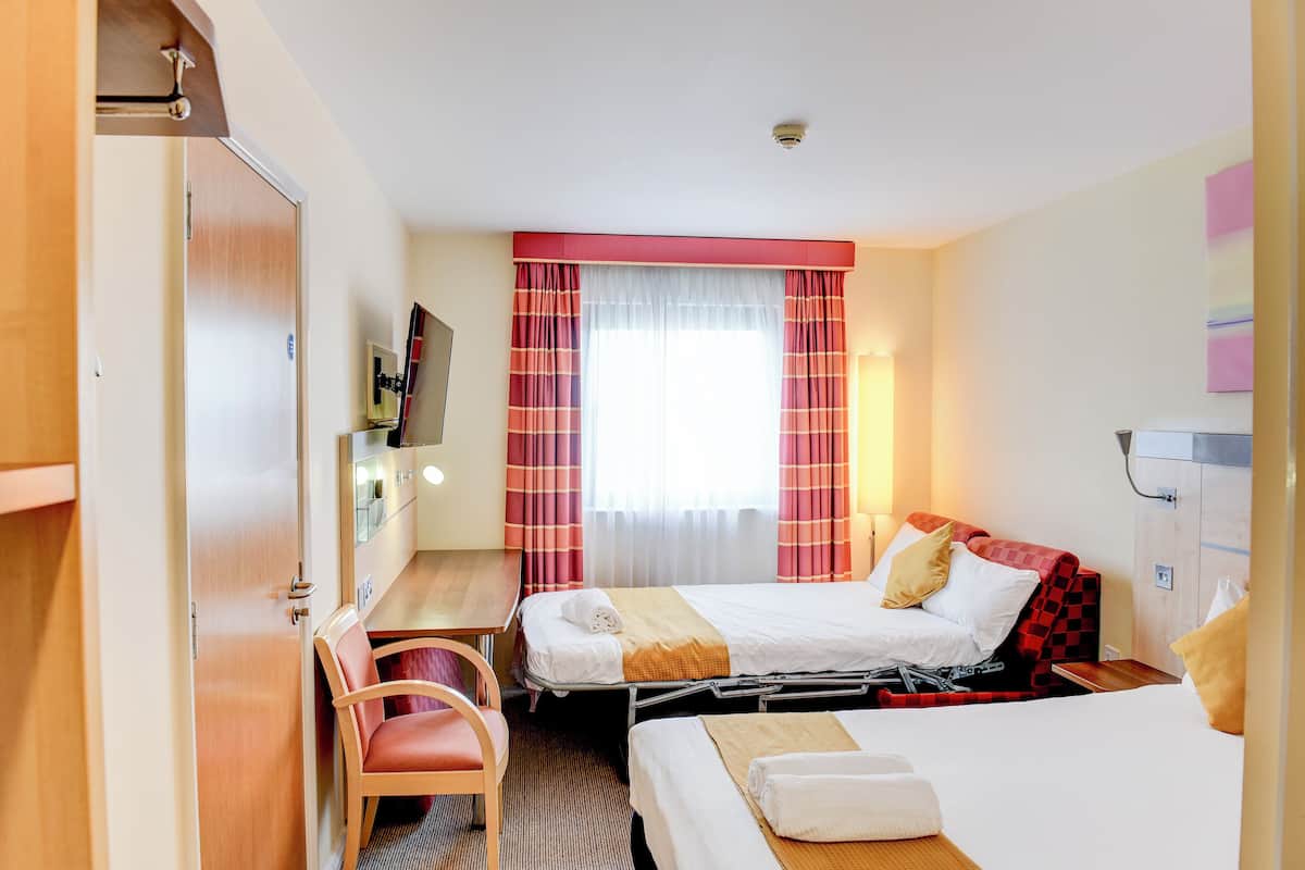 Quarto de hotel com duas camas, uma mesa, uma cadeira, uma televisão e uma janela com cortinas.