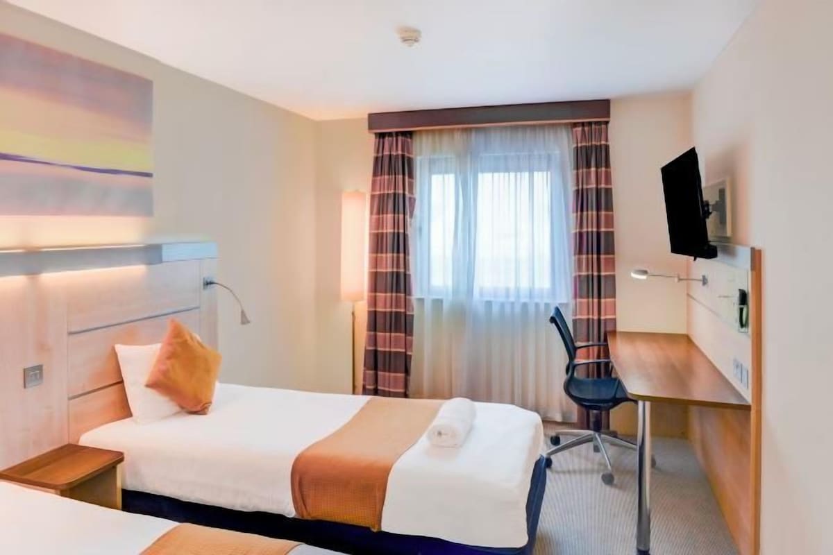 A hotel room with a bed, a desk, a chair, a TV, and a window with curtains.