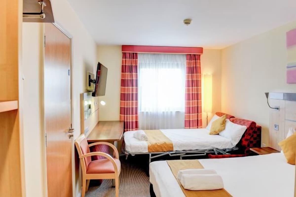 A hotel room with two beds, a desk, a chair, a TV, and a window with curtains.