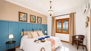 Un dormitorio con cama, mesita de noche, una silla, ventana con cortinas y una obra de arte en la pared.