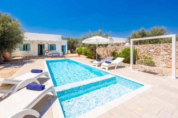 A swimming pool with lounge chairs and umbrellas, surrounded by a stone wall and vegetation.