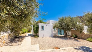 A white building with a small window, surrounded by olive trees and a gravel path leading to a paved area.