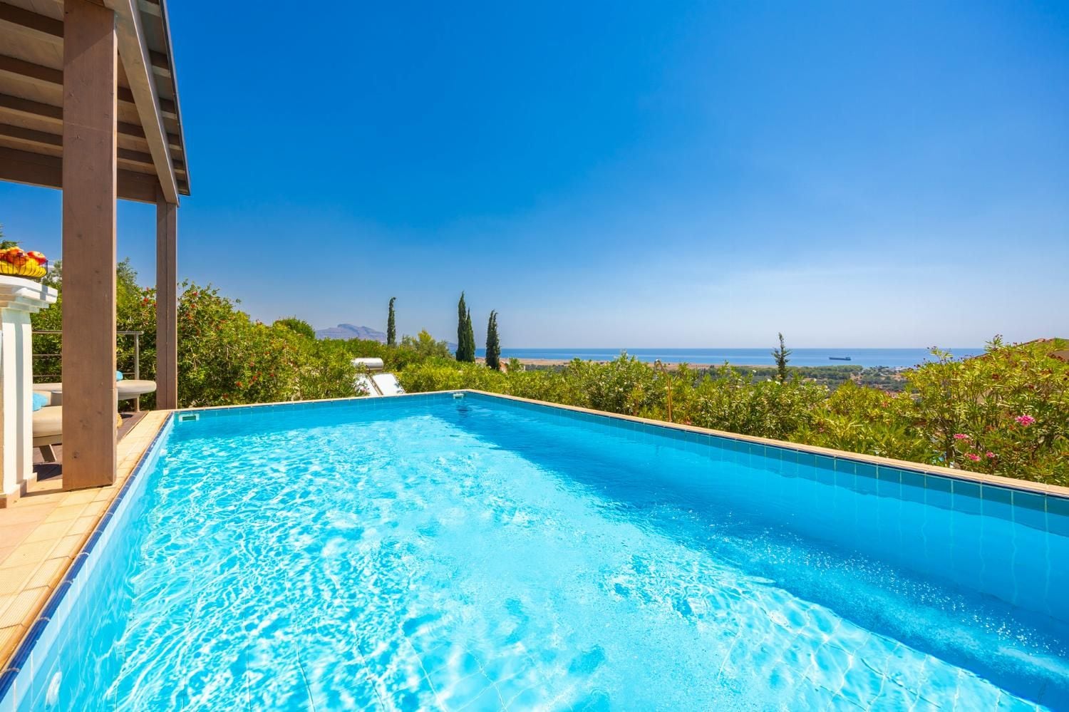 Une piscine rectangulaire à l’eau d’un bleu limpide, entourée de verdure et offrant une vue sur la mer.
