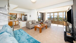 A spacious living area with a sofa, coffee table, and a television. There is a kitchen in the background and a balcony with a view. - Villa Daphne - Two Bedroom Villa, Sleeps 4 (Kalathos)