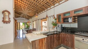 A modern kitchen with wooden cabinetry, a central island, stainless steel appliances, pendant lighting, and a view into the dining area. - Residência Flora (Porto Seguro)