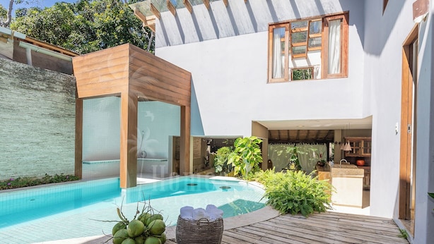 A modern outdoor pool area with a wooden deck, a glass-enclosed spa, and lush greenery. - Residência Flora (Porto Seguro)