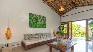 A spacious living area with a wooden ceiling, a large green tropical-themed painting, a glass coffee table, and a cushioned bench. - Residência Flora (Porto Seguro)
