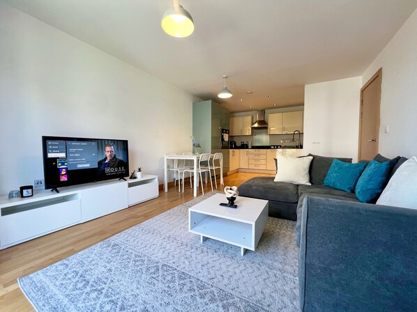 Een moderne woonkamer met een flatscreen televisie, een witte salontafel, een grijze bank en een keuken op de achtergrond.