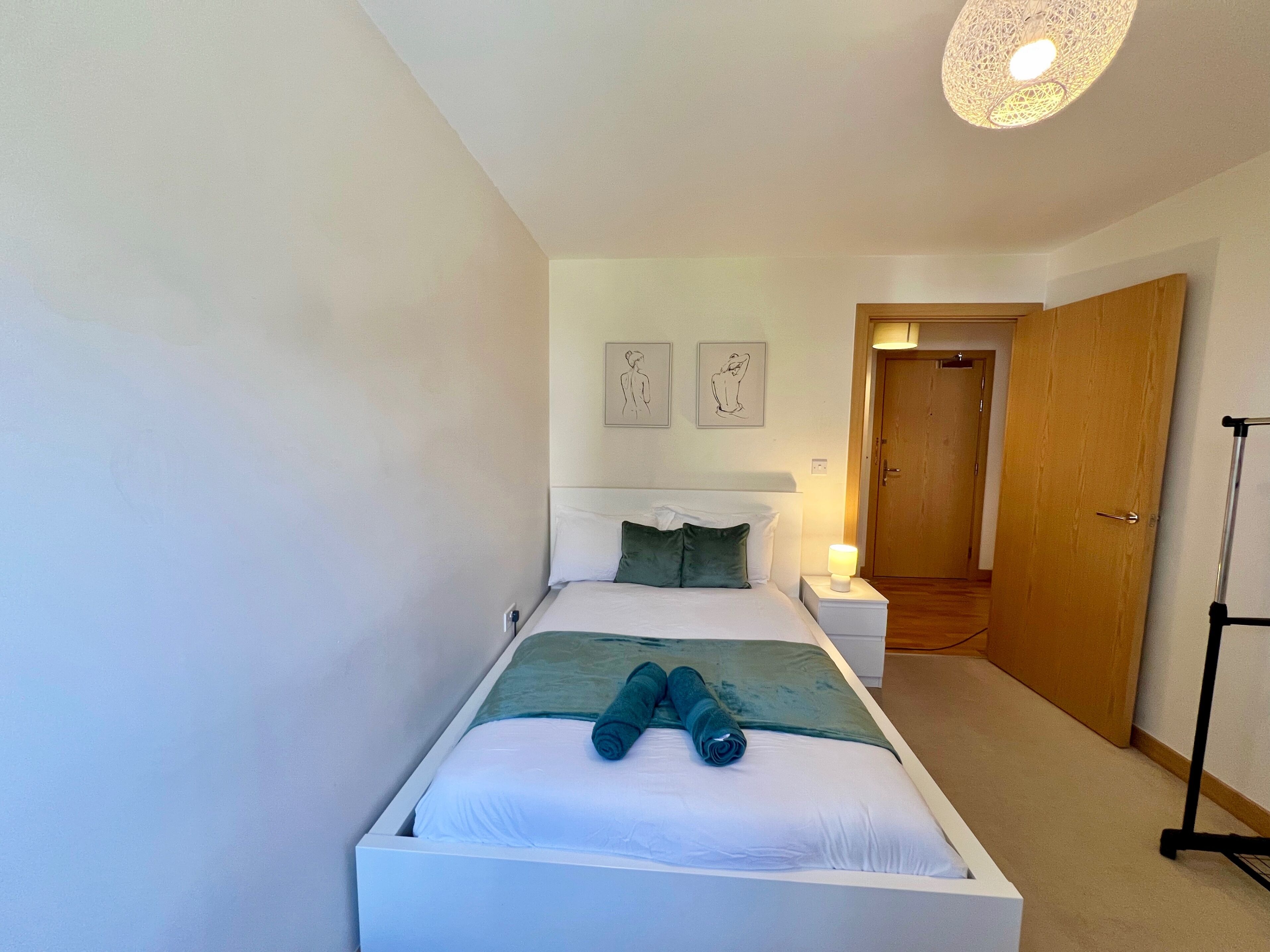Un lit bien fait, avec une tête de lit blanche, un plaid vert et des serviettes enroulées. On peut voir deux tables de chevet avec des lampes, et une porte en bois à l’arrière-plan.