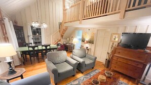 Ein geräumiges Wohnzimmer mit hoher Decke, einer Holztreppe und einem Essbereich mit einem grünen Tisch und Stühlen.