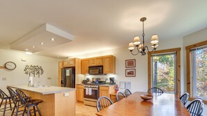 A modern kitchen with wooden cabinets, a central island with bar stools, a dining table with chairs, and a chandelier. - Ski-In/Ski-Out at Jay Peak Resort Condo! (Jay)