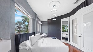 A modern bedroom with a large bed, a bedside table, a mirror, and a view of palm trees. - Villa Tortuga: 10 BR 9,500 sq. ft Extravagant Miami Estate w pool, movie theater (Miami)