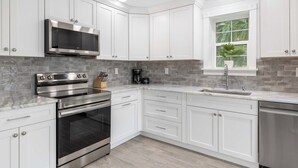 A modern kitchen with white cabinetry, stainless steel appliances, a large window with a plant, and a tiled backsplash. - Water side Upstairs Condo in Sebastian FL (Sebastian)