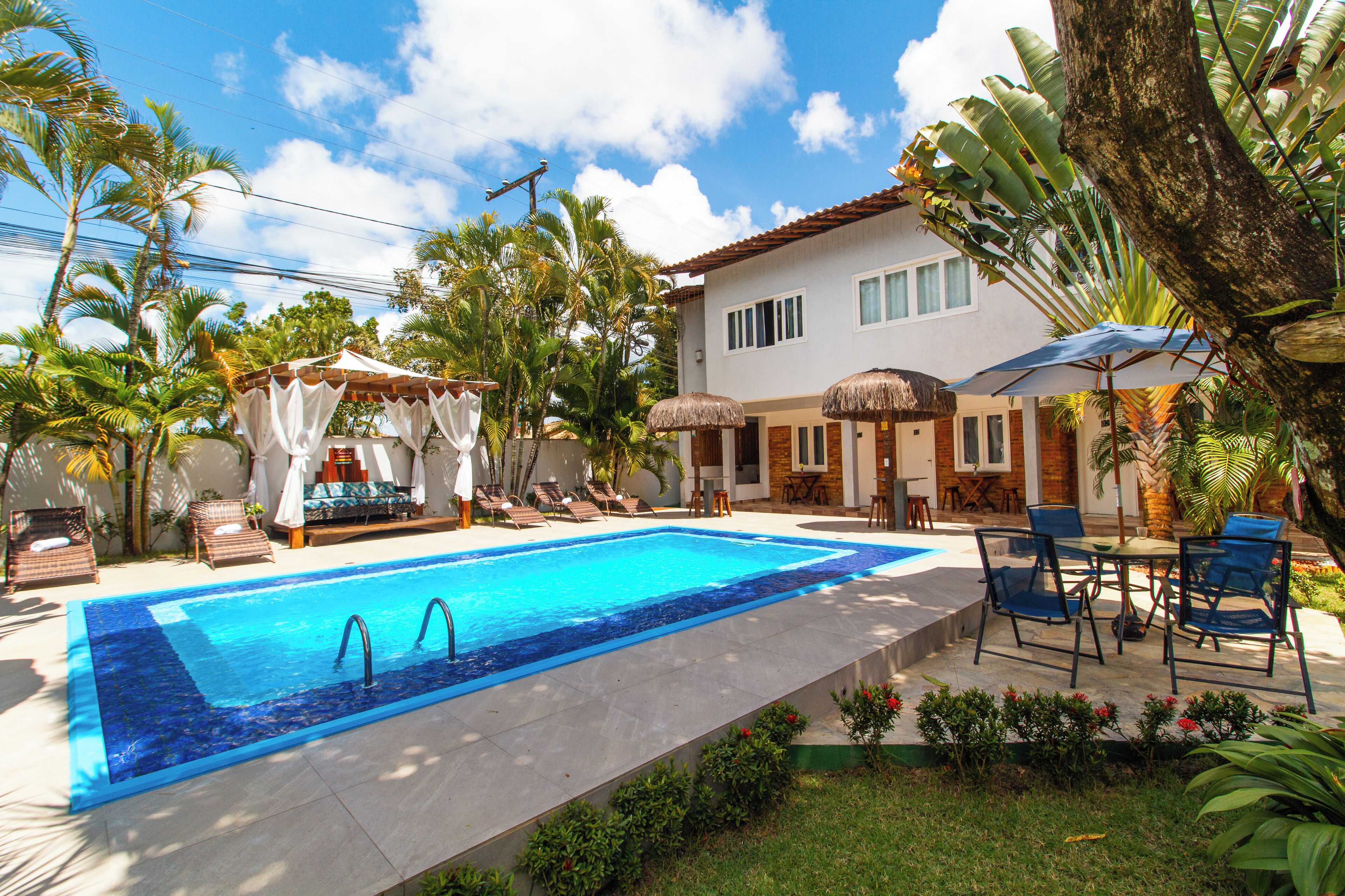 A swimming pool with lounge chairs and umbrellas, surrounded by tropical plants and a house.