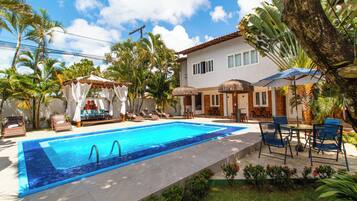 A swimming pool with lounge chairs and umbrellas, surrounded by tropical plants and a house.