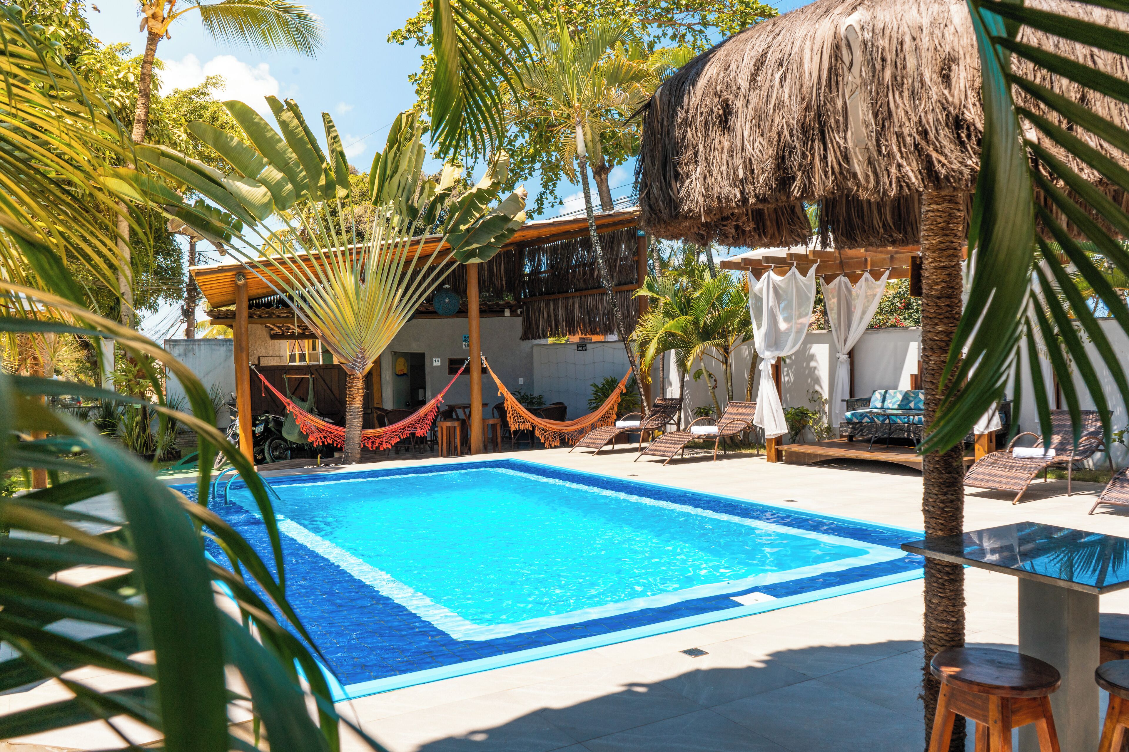 Una piscina con sillas de descanso y una hamaca, rodeada de palmeras y una estructura de techo de paja.