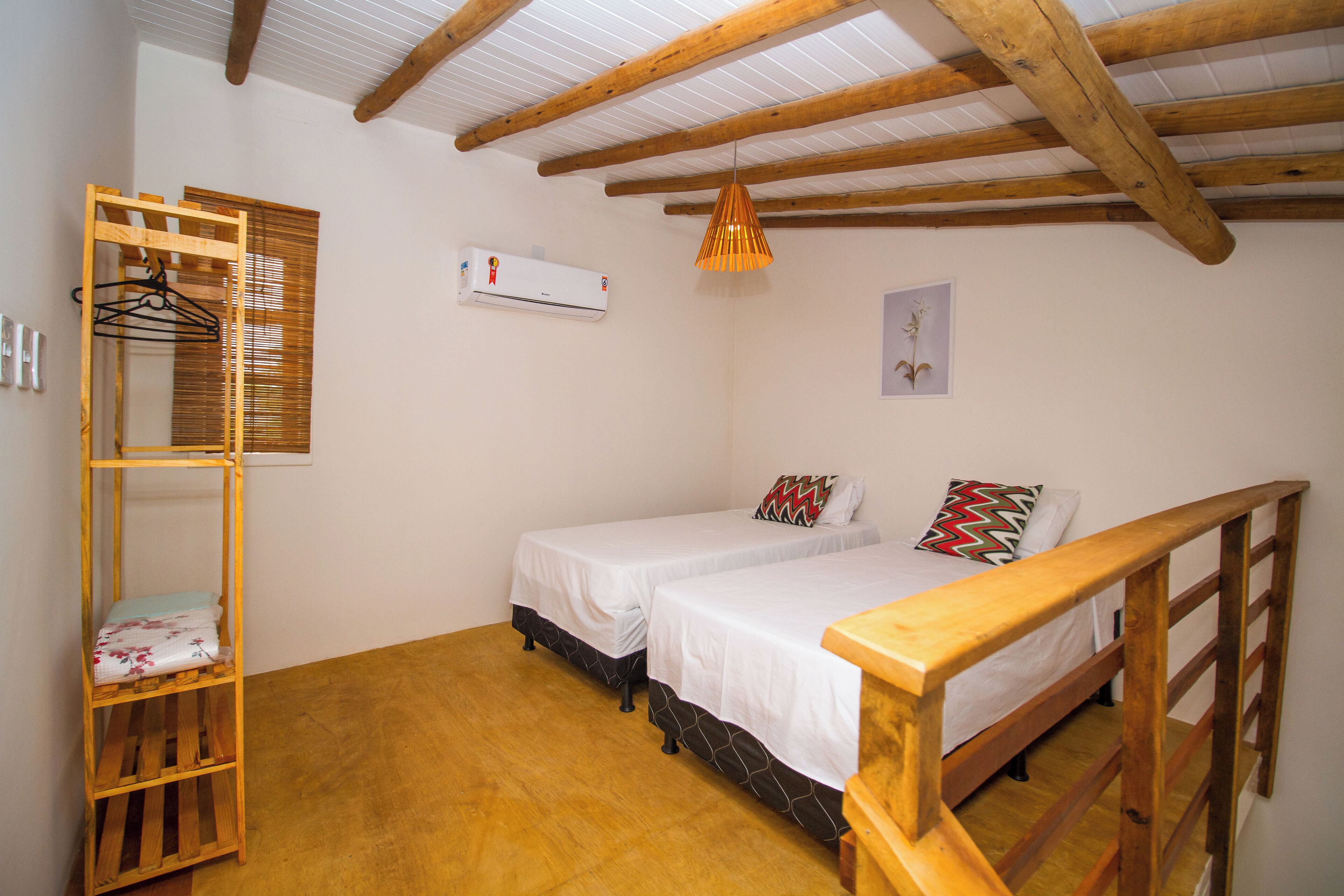 Une petite pièce bien éclairée, avec des poutres en bois au plafond, deux lits simples avec du linge de lit blanc, une étagère en bois avec un porte-vêtements et un appareil de climatisation fixé au mur.