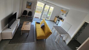 A modern living room with a sofa, a coffee table, and a TV. There is a kitchen area with a sink and cabinets. The room has a sliding glass door leading to a balcony. - Duplex apartment Mimizan plage (Mimizan)