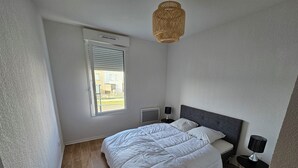 A bedroom with a large bed, a window with blinds, a ceiling light fixture, and a bedside table with a lamp. - Duplex apartment Mimizan plage (Mimizan)