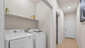 Een wasserette met twee witte Samsung wasmachines en drogers, een plank met schoonmaakmiddelen en een deur naar een andere ruimte.