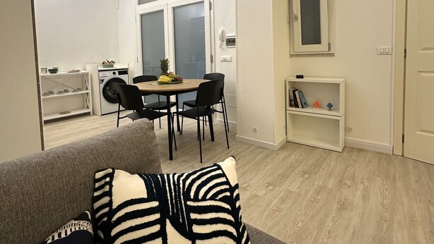 Ein modernes Wohnzimmer mit einem grauen Sofa, einem Essbereich mit rundem Tisch und Stühlen sowie einer Küche im Hintergrund.
