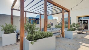 An outdoor patio area with wooden pergolas, white planters with greenery, and a seating area with a sofa and chairs. - Six Flags/The Rim - Enchanting Modern Studio, King (San Antonio)