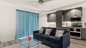 A modern living room with a dark blue sofa, glass coffee table, and a ceiling fan. - Six Flags/The Rim - Enchanting Modern Studio, King (San Antonio)