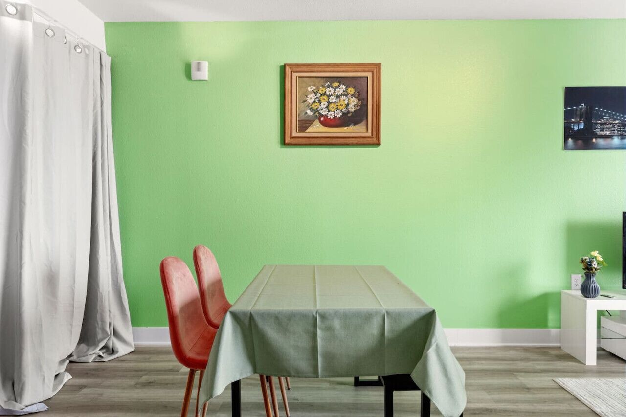 En matplats med ett bord täckt av en grön duk, två röda stolar, ett vitt sidobord med en vas och en inramad tavla med blommor på väggen.