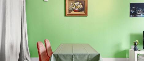 En matplats med ett bord täckt av en grön duk, två röda stolar, ett vitt sidobord med en vas och en inramad tavla med blommor på väggen.