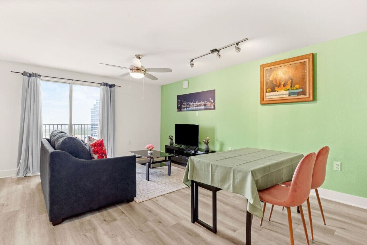 Un salon moderne avec un canapé, une table à manger et des chaises. On y trouve un téléviseur à écran plat posé sur un meuble, un ventilateur de plafond et un tableau accroché au mur.