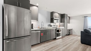 A modern kitchen with stainless steel appliances, grey cabinetry, and a grey sofa in the living area. - Six Flags/The Rim - Chic Modern Studio, King Bed (San Antonio)