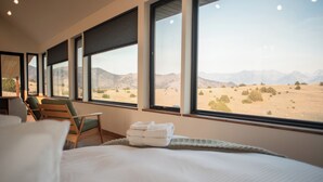 A hotel room with a large bed, a chair, and a view of a desert landscape through large windows.