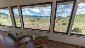 A room with large windows offering a view of a mountainous landscape. There are two wooden chairs with cushions and a small round table.