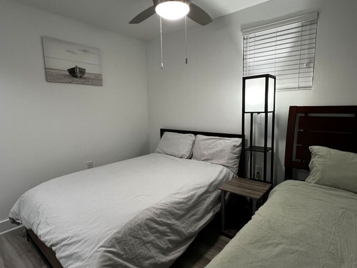 A bedroom with a single bed, a nightstand, a ceiling fan, a window with blinds, and a framed picture on the wall.