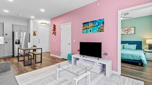 A modern living room with a pink accent wall, a flat-screen TV, a glass coffee table, and a dining area with a white table and chairs.