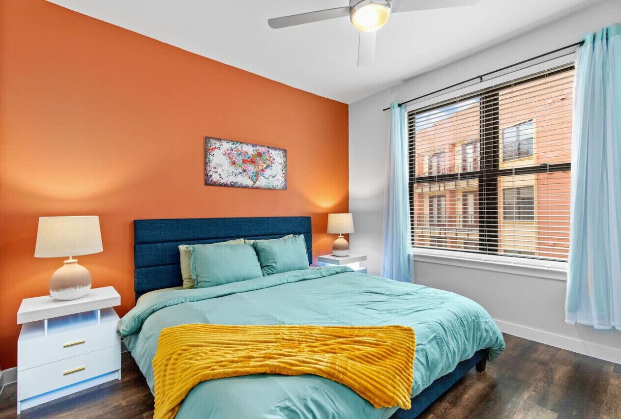 Quarto com cama, mesas de cabeceira, ventilador no teto, janela com persianas e vista para a cidade.