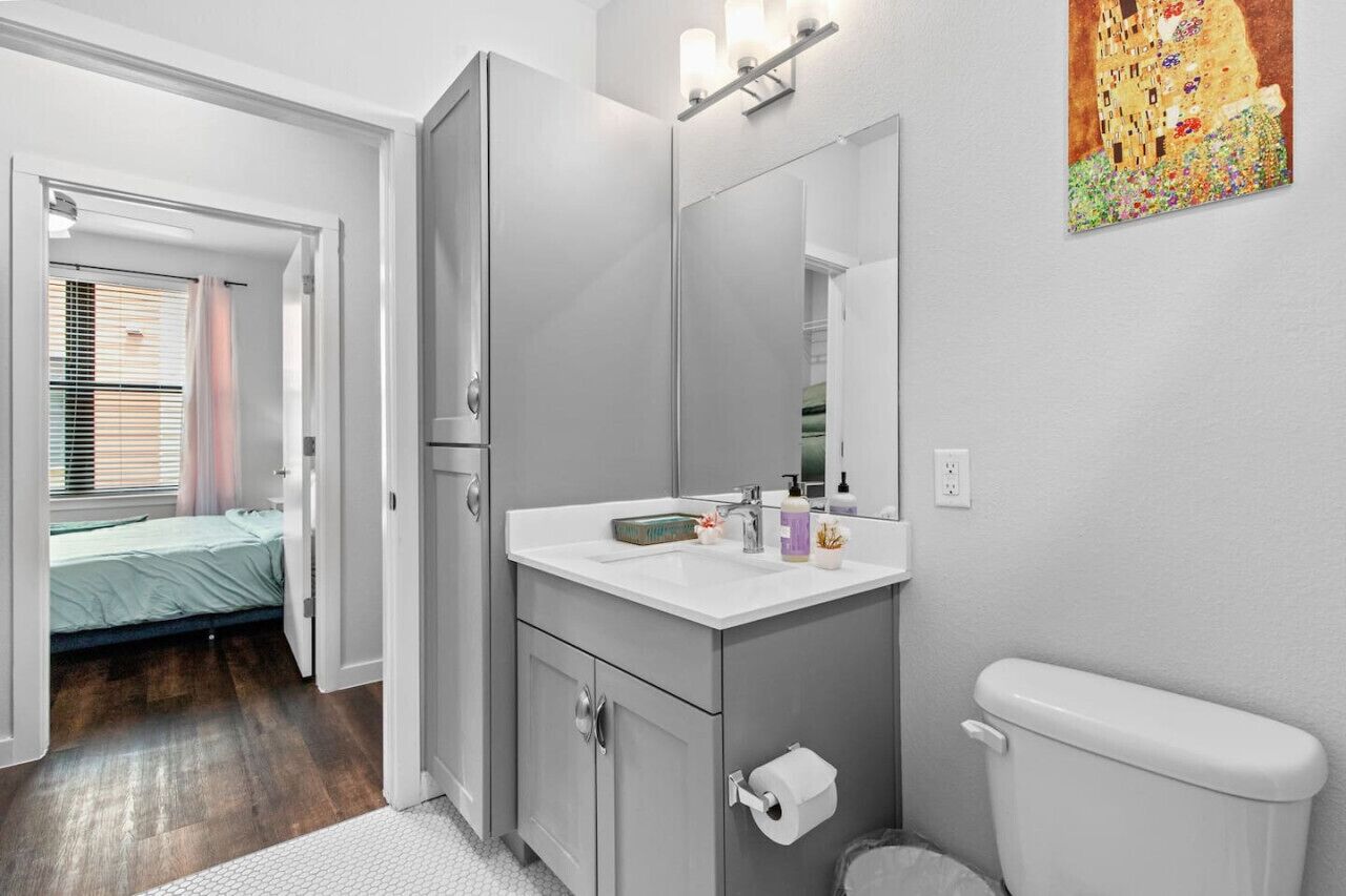 A modern bathroom with a large mirror, a white sink, and a toilet. There is a bed visible in the adjacent room.