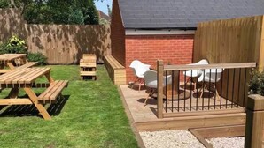 Un jardin avec une terrasse en bois, des tables de pique-nique et des chaises blanches.