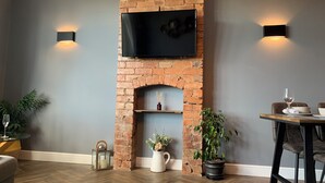 Une pièce avec une cheminée en briques, un téléviseur fixé au mur, deux lampes murales, une table à manger avec des chaises et une plante en pot.