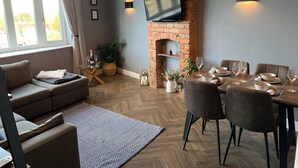 Un salon moderne avec une cheminée en briques, un téléviseur à écran plat, un coin repas avec une table et des chaises en bois, et une grande fenêtre avec des rideaux.