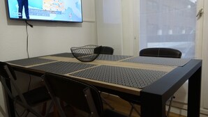 Een eettafel met een zwarte draadmand, zwarte stoelen en een televisie aan de muur met een weersvoorspelling.