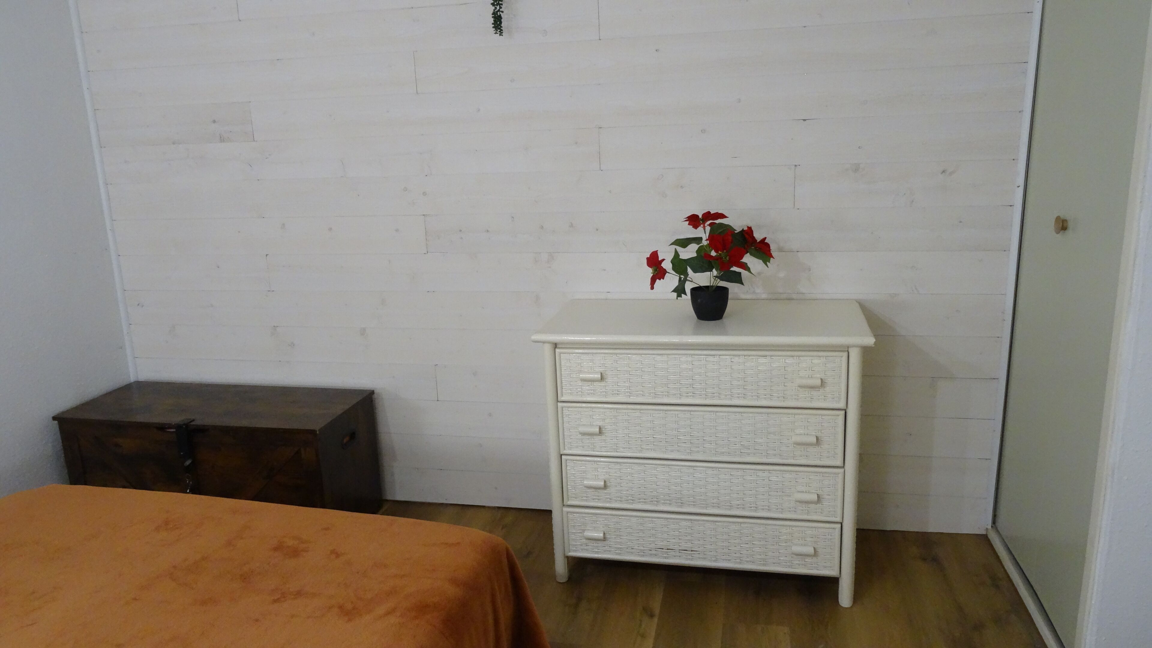 A room with a white dresser, a wooden chest, and a bed with an orange bedspread.