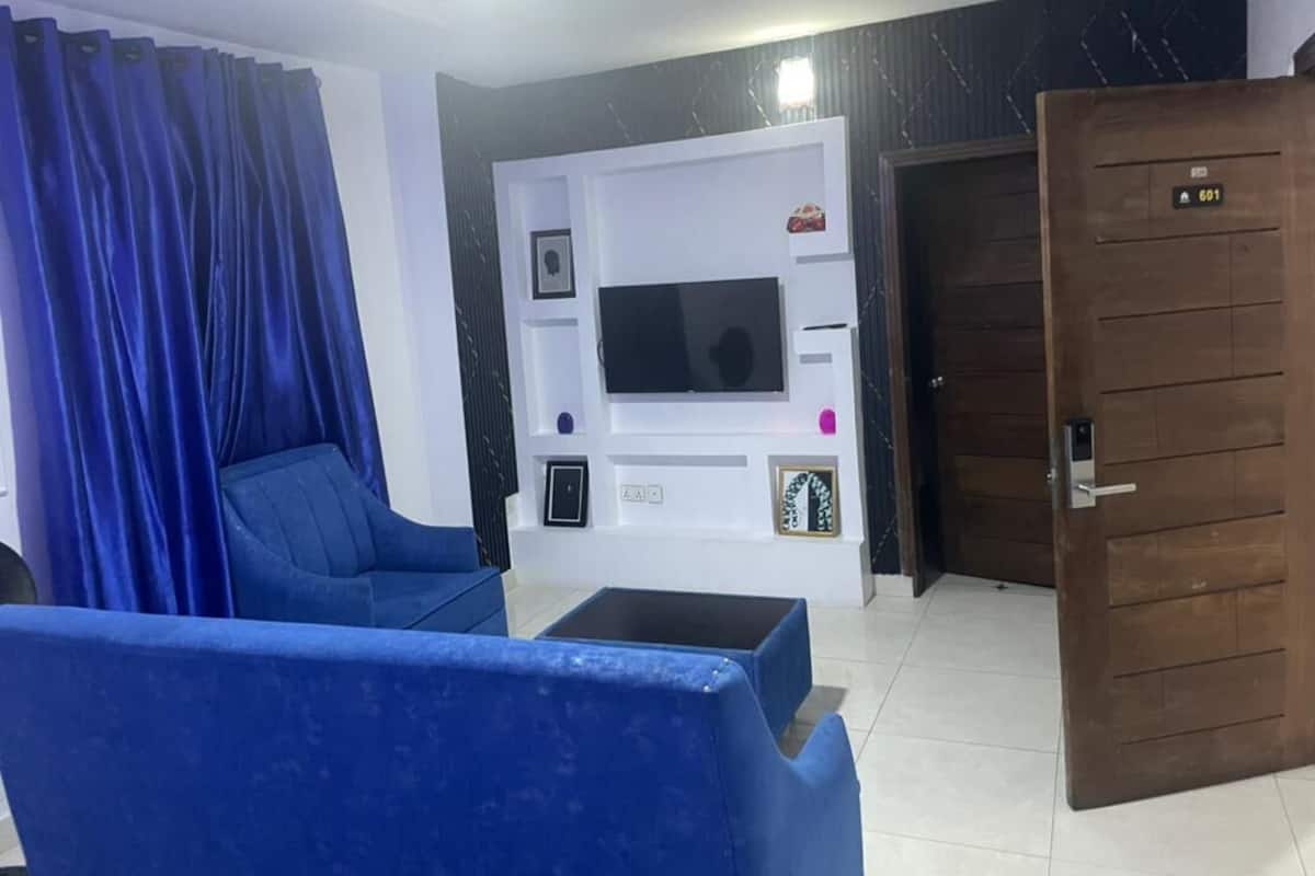 Una sala de estar con un sofá azul, un televisor instalado en una pared blanca y una puerta de madera con una placa de número.